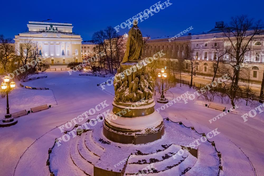 На фотографии изображён памятник Екатерине II, установленный в центре площади Островского. На заднем плане возвышается фасад Александринского театра — одного из старейших театров России, созданного по проекту архитектора Карла Росси. Снежный покров и мягкое освещение придают сцене сказочную атмосферу. Улицы пустынны, что подчёркивает спокойствие зимнего вечера. Всё пространство оформлено праздничной подсветкой к новогодним праздникам.